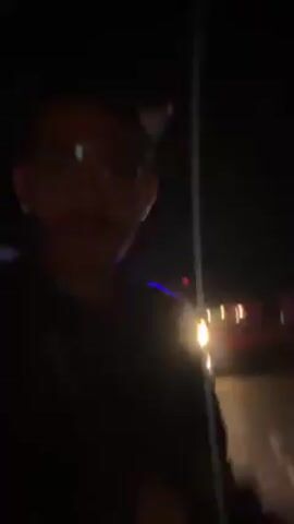 Here is a concise description of the video:

The video shows a person holding a camera and walking down a dark street at night. They are wearing glasses and a dark jacket. There are vehicles visible in the background, including one with flashing blue lights. The lighting is poor, making it difficult to see details clearly. The focus is primarily on the person holding the camera, with the background elements appearing blurry and indistinct.