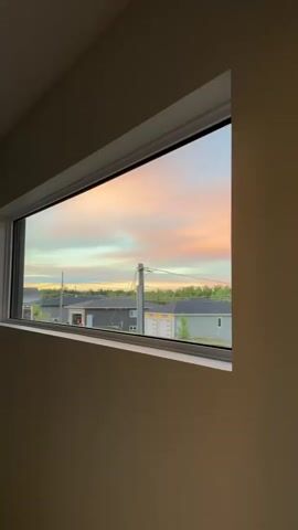 A horizontal, rectangular window frames a view of a suburban neighborhood during what appears to be dusk. The sky above is painted with soft pastel hues of pink, orange, yellow, and blue, indicating a sunset or sunrise. Below the sky, a line of houses with modern architecture is visible, featuring pitched roofs and garage doors. Some houses are light-colored with stone accents, while others are darker gray. The houses are surrounded by neatly manicured green lawns. Utility poles and power lines crisscross the scene, a common element in residential areas. The overall environment suggests a peaceful and tranquil setting. The video captures a static view through the window with no movement or activity depicted.