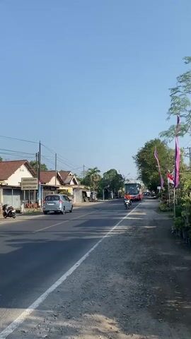 A daytime, vertical, outdoor shot shows a road with vehicles and buildings. On the left side of the road are low-rise buildings with white walls and brown roofs, some with signage. A silver car is parked in front of one building. A motorcycle is parked on the side of the road. A paved road runs through the center of the frame, with a white line marking the lane. On the right side of the road is a dirt shoulder with plants and trees. Two purple flags and one red and white flag are visible on poles. A large white and red bus is driving down the road. A person on a motorcycle, wearing an orange shirt, is also driving on the road, passing the bus. The sky is clear and blue.