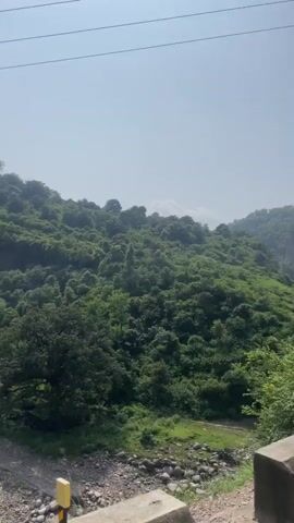 The video shows a view from a vehicle moving along a road, likely in a hilly or mountainous region. Dense green vegetation covers the hillsides, suggesting a natural, possibly rural environment. A concrete barrier is visible on the right side of the road, a common roadside safety feature. As the vehicle continues, a pink temple with traditional architectural elements, including a spire, comes into view. A yellow directional sign indicating a curve to the right is also visible next to utility poles. The scene appears to be peaceful and scenic, with a clear sky overhead.