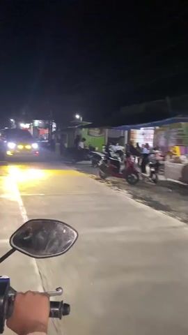 The video captures a nighttime scene from the perspective of someone riding a motorcycle. The motorcycle's mirror is visible in the foreground, and the road ahead is illuminated by streetlights and the headlights of oncoming vehicles. In the first few seconds, a silver car with bright yellow headlights passes by, its beams intensely illuminating the road. The background features a street lined with small shops and buildings, some of which are lit up. Several parked motorcycles and scooters are visible along the roadside. People can be seen in the distance, suggesting a populated area. The overall environment appears to be a quiet street in a town or village, with businesses open and activity present despite the late hour. The camera appears to be handheld and experiences some movement consistent with riding a motorcycle.