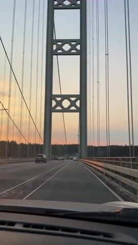 The video shows a view from inside a vehicle driving across a suspension bridge during sunset. The camera is positioned to show the dashboard and windshield, with the bridge's suspension cables and a large tower visible ahead. Several cars are driving in the same direction on the bridge. The sky displays a gradient of orange and yellow hues from the setting sun, with some clouds present. A line of trees can be seen in the distance beyond the bridge.