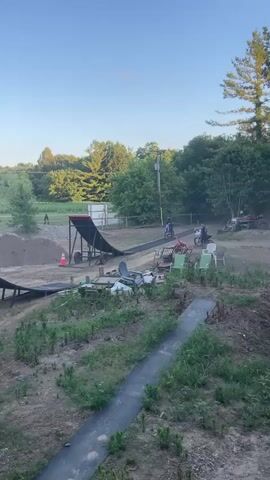 A motocross rider is captured in mid-air after launching off a ramp. The rider and their blue and white dirt bike are silhouetted against a clear blue sky. Below the airborne bike, a large inflatable ramp is visible, designed for riders to land on. The surrounding environment appears to be an outdoor recreational area, possibly a backyard or private track, with trees and foliage in the background. Another dirt bike is visible on the ground near the ramp. The video transitions to the rider landing on the inflatable ramp, with a soft landing visible.