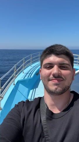 A man is taking a selfie on a boat. He is wearing a black t-shirt and has dark hair and a beard. The boat is blue and white and is on the ocean. The sky is clear and blue.