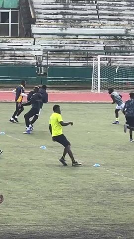 A group of men are on a grassy field, likely a football pitch, practicing. In the foreground, a man in a neon yellow shirt and black shorts runs past blue cones. To his right, a man in black shorts and a black shirt is running away from the camera. To the left, a man in a black jacket and black pants runs past. In the mid-ground, there are several other men. One man in a blue and white jersey runs past a man in a grey shirt and black shorts. Another man in a dark blue shirt and black shorts runs towards the right. Behind them, a man in a black shirt runs past. Further in the background, a football goal with a net is visible. The stands of a stadium, with weathered white and green seating, are behind the field. The sky appears overcast.