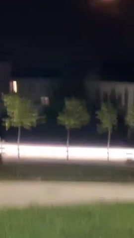 A video captures a full moon with an orange hue in a dark sky. The moon is partially obscured by thin clouds. Below the moon, silhouetted buildings are visible, with rows of green trees in front of them. A bright, horizontal light band runs across the middle ground, suggesting streetlights or vehicle headlights. The foreground shows a paved surface and some grass. The camera appears to be handheld and moves slightly throughout the short clip.