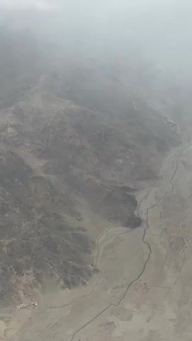 The video shows an aerial view of a mountainous, arid landscape. The terrain is characterized by rugged slopes, deep ravines, and a dry riverbed snaking through the valley. The colors are predominantly muted browns and grays, indicating sparse vegetation. The sky is overcast with thick clouds, creating a hazy and diffused light. There are no visible people or vehicles in the frame, only the natural features of the land. In the lower left corner, a small cluster of buildings can be faintly discerned, suggesting a sparsely populated area. The overall impression is one of a remote and harsh environment.