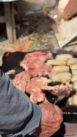 A person's hands are shown in the foreground, cooking meat on a flat, dark surface, possibly a grill or griddle. The meat is arranged in rows, with some pieces appearing pink and raw, while others are starting to brown. There is smoke and flame visible around the meat, indicating it is cooking. The background is blurred but appears to be an outdoor setting with some debris and a white tarp. The person's hands are aged and wrinkled, and they are wearing a grey long-sleeved shirt. The focus is on the cooking process and the food.