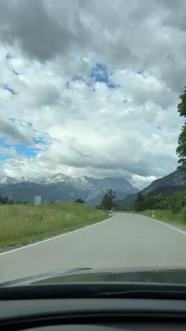 The video shows a view from the inside of a car driving on a paved road. The road curves to the right. On the left side of the road, there is a grassy field with tall grass and some distant trees. Beyond the field, a range of snow-capped mountains is visible under a cloudy sky. There are some traffic signs and a guardrail along the side of the road. On the right side of the road, there are trees and a hillside. The weather appears to be overcast with patches of blue sky.