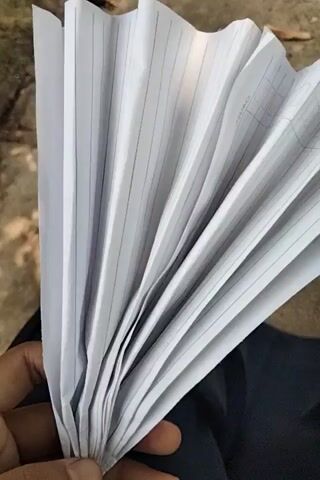 The video shows a person's hand holding a fan of lined paper. The paper appears to be from a notebook or a ledger, with horizontal lines and some vertical columns visible. The background is blurry but appears to be outdoors, with some leaves and dirt on the ground. The hand is holding the papers in a way that suggests they might be about to be handed over or reviewed. The lighting is natural, indicating it's daytime. No faces or other identifiable features of the person are visible.