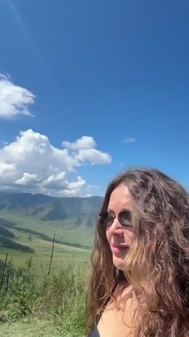 The video shows a woman with long, wavy brown hair and sunglasses looking at the camera. She is standing outdoors with a landscape of green hills and mountains in the background. The sky is blue with white clouds. In the foreground, there is a muddy puddle. The camera appears to be held by the woman, suggesting a selfie video. The woman is smiling slightly. The video is short, lasting only a few seconds.
