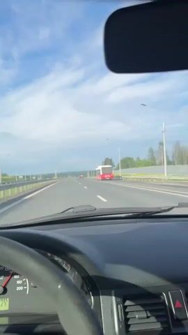 A first-person point-of-view video shows a person driving a Lada car on a multi-lane highway during the day. The sky is bright with some clouds. The driver's hands are visible on the steering wheel. The dashboard shows the speedometer indicating a speed between 100-110 km/h and the odometer displaying 199995 km. An infotainment system with "Bluetooth" visible is on the center console. Through the windshield, a red bus is seen driving in an adjacent lane on the right. The road extends straight ahead with streetlights and some greenery along the sides. The setting is consistent with Ust'-Syny, Russia.