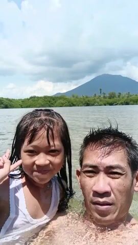 A man and a young girl stand waist-deep in water, facing the camera. The girl has wet hair and is wearing a light-colored top. She makes various silly faces. The man, also with wet hair, remains relatively stoic, looking into the camera. Behind them is a body of water bordered by green trees, with a mountain visible in the distance under a cloudy sky.