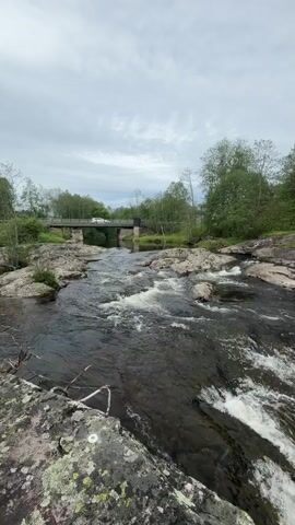 The video shows a rocky river flowing over a short waterfall or rapids. The water is dark and churning, with white foam forming as it rushes over the rocks. Lush green trees line both sides of the river. A small bridge is visible in the distance spanning the river. The sky is overcast with grey clouds. No people are visible.