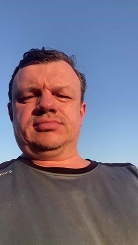 A man is shown in a selfie video. He is wearing a gray t-shirt. The background changes between a clear blue sky, a landscape with buildings, and a vineyard.