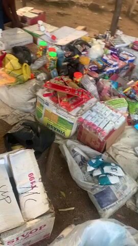 The video depicts an outdoor market scene, likely in Reak, Nigeria. The foreground is filled with a variety of goods for sale, predominantly stacked in a chaotic but organized manner. These include numerous green packets, possibly detergent or soap, many with "Viva" branding visible. There are also boxes containing smaller items, such as what appear to be bars of soap or packaged food items, some in red and white packaging. Other objects include plastic bags, a large white sack with text and a recycling symbol, and what seems to be a stack of books or notebooks.

In the background, under a thatched roof structure supported by wooden poles, several people are gathered. They appear to be interacting, possibly as vendors and customers. A person in a bright pink shirt is visible in the leftmost part of the frame, bent over a table displaying items. Other individuals are standing and talking. A motorcycle is parked behind the group.

The setting appears to be a somewhat dusty, outdoor environment with sparse vegetation and large rocks in the distance, suggesting a semi-arid or rocky landscape. The lighting indicates it is daytime, with sunlight casting shadows, possibly late afternoon given the warm tones. The overall atmosphere seems to be that of a bustling local marketplace. There are no strong emotional cues immediately apparent, but the activity suggests commerce and social interaction. Visible text includes "Viva" on the green packets and some text on the white sack, though much of it is illegible.