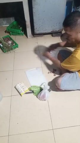 A man in a yellow shirt and grey shorts kneels on a tiled floor, chopping ingredients. He is surrounded by food items, including eggs, onions, and leafy greens. Two packages of cookies are also visible. In the background, green crates hold more produce. A large, metal appliance, possibly a refrigerator or cabinet, stands against the wall. The lighting is dim.