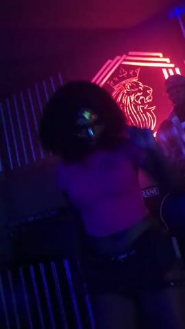A person with dark curly hair wearing a pink crop top and black shorts dances in a club. Neon lights in red and purple illuminate the scene, with a lion logo displayed on the back wall. A person wearing a baseball cap and plaid shirt can be seen in the background behind a DJ setup. The dancer has glitter makeup on their face and is moving their arms and body to the music.