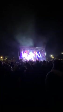 A person recorded a band performing in front of a large crowd at night. The video starts with a view of the crowd in silhouette, then zooms in on the stage where the band is performing. The stage is lit with bright white, red, and blue spotlights. The band includes multiple singers and musicians. The singers are standing front and center on the stage. The crowd is standing and watching the performance. There is smoke rising into the air around the stage.
