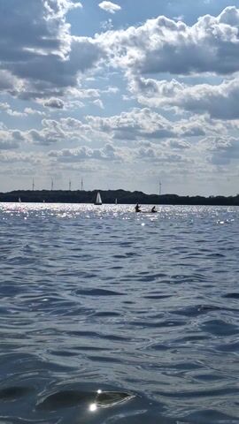 The video shows a wide shot of a body of water with a cloudy sky above. The water is choppy and reflects the sunlight. In the distance, there is a shoreline with trees and what appear to be wind turbines. There are a few sailboats on the water and two figures in the water who appear to be swimming or wading. The image is rotated 90 degrees counterclockwise.