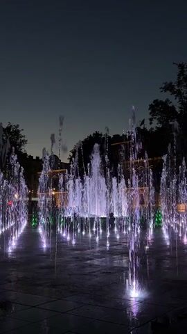 The video shows a fountain at night. The fountain has multiple jets of water that are lit up with changing colors. In the background, there is a building with lights on and trees. The sky is dark. The fountain is in a paved area, possibly a plaza or park. The water jets are arranged in rows and around a central, larger jet. The colors change from white and green to purple and blue.