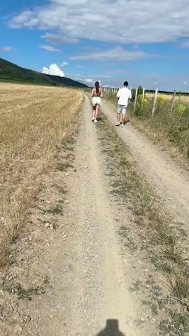 A man and woman walk down a dirt path in a rural landscape. The path is bordered by a dry, golden field on the left and a fence with tall grass on the right. In the background, a lush green mountain rises under a bright blue sky dotted with white clouds. The scene conveys a sense of peaceful exploration and natural beauty.
