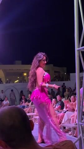 In an outdoor evening setting, a woman performs a lively belly dance on an illuminated stage. She wears a vibrant, two-piece pink costume adorned with sequins, fringes, and flowing sheer fabric, with her long, dark hair flowing freely. The stage is bathed in purple light. Around the stage, numerous audience members are seated at tables and benches, watching the performance. Many are smiling and some are clapping, indicating enjoyment. In the background, white buildings with arches are visible under the dark night sky, suggesting a resort or entertainment complex, possibly in Egypt based on the provided context. A metal scaffolding structure stands on the right side of the frame. The dancer moves energetically, engaging with the audience.