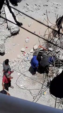 From a high vantage point, the video shows several people standing around a debris-filled area with exposed rebar. The ground is light colored, and there are small pieces of rubble scattered everywhere. A man in a blue shirt is crouched down, while others stand nearby or lean over, seemingly inspecting or trying to assist someone amidst the twisted metal. There are reddish stains on the ground near the crouched man, suggesting potential injuries. The environment appears to be a construction site or a collapsed structure, and the people seem to be responding to some kind of accident or incident.