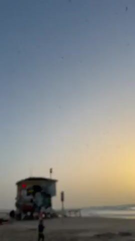 The video shows a beach scene at sunset. A lifeguard tower stands on the sand, covered with graffiti. A large flock of birds flies in the sky above the beach. People are visible on the beach: a child walks near the lifeguard tower, a man walks by, and a person reclines in a beach chair. Tall buildings are visible in the background. The sky is blue with a gradient of orange near the horizon.