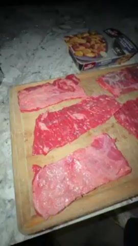 The video shows a person preparing food on a countertop. There are several cuts of raw beef on a wooden cutting board and a metal tray. The person is seen touching the meat. A package of roasted potatoes and a container of sliced strawberries are visible on the countertop. The countertop has a speckled granite-like surface. The lighting in the video is dim.