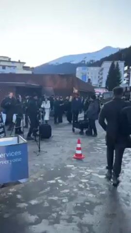 The video shows a crowd of people, including individuals who appear to be members of the media with cameras and tripods, gathered on a paved area. A security detail is also present. The location appears to be a town or village, with buildings visible in the background. A snow-capped mountain is visible in the distance. There is an orange and white striped traffic cone is visible in the foreground. The general atmosphere suggests a public event or media gathering.