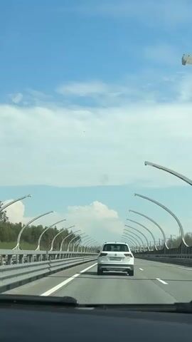 The video shows a car's perspective while driving on a highway with a white car in front. The sky is blue with scattered white clouds. The road is lined with a guardrail and unique street lights with curved, arching arms. Trees are visible on the side of the road. The car's dashboard is partially visible in the bottom of the frame.