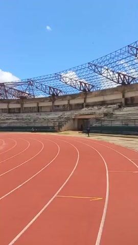 The video shows an outdoor athletic stadium. In the foreground is a red running track with white lines marking the lanes. In the middle ground, a grassy field is visible, likely a football pitch, with a corner flag on the right side. In the background, the stadium seating is visible, appearing somewhat dilapidated with scaffolding on some sections. The sky is blue with scattered white clouds. One person is briefly visible standing on the running track in the middle distance. Another person is seen from behind on the track in the foreground, holding up a phone, possibly taking a picture or video. The overall impression is of an aging or underutilized sports facility.