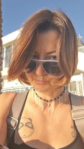 The video shows a woman with short, brown hair and fair skin standing outdoors on a sunny day. She is wearing aviator sunglasses with a light blue frame. Beneath the sunglasses, blue eyeliner is visible. She has a nose ring and is wearing a black and silver beaded choker necklace with dangling charms. She has a black tank top and a backpack. A tattoo of a panda’s face is visible on her chest. She raises her hand to her hair, then removes her sunglasses to look directly at the camera. A palm tree and a white building can be seen in the background.