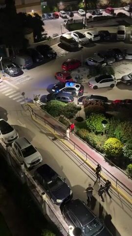 The video, shot from an elevated angle at night, captures a parking lot scene with a visible police presence. Multiple cars, some parked and some moving, are observed, including a Spanish police vehicle with flashing blue lights. Several individuals, seemingly police officers dressed in dark clothing, are seen running across a crosswalk and along a walkway. A couple, one in a red shirt and the other in white, are also seen walking on the ramp. The parking lot itself is surrounded by buildings and greenery, suggesting an urban or suburban environment. The camera remains stationary throughout the video, offering a consistent overview of the unfolding activity.