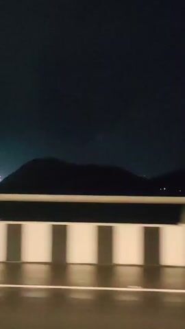 The video shows a nighttime view from a moving vehicle. In the foreground, a wet asphalt road and a white barrier with square posts are visible, blurred by motion. Beyond the barrier, a dark, silhouetted mountain range occupies the middle ground. In the distance, the lights of a city or industrial area twinkle. The sky is dark and cloudy.