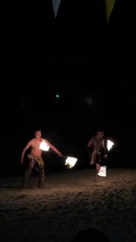 Two men in short skirts are performing a fire dance on a beach at night. They are holding flaming objects in their hands, which they spin and throw. There are a few people watching in the background. There are colorful triangular flags hanging above them.