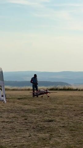 A man is standing on a grassy field with a remote-controlled airplane in front of him. The man is holding a remote control in his hands and appears to be operating the airplane. The airplane is red and white. The field is dry and the grass is short. In the background, there are rolling hills and a hazy sky. The man is wearing a blue t-shirt and shorts. He has a baseball cap on his head. The airplane is on its landing gear. The man appears to be focused on the airplane.