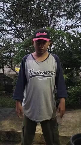 A man wearing a gray, long-sleeved shirt with blue sleeves, olive green cargo pants, and a black and pink baseball cap is standing in a garden area. He is in the foreground and is facing the camera. The man is performing a dance-like motion with his arms and body. Behind the man are various plants, trees, and a glimpse of a building in the background. The scene appears to be outdoors, possibly in a backyard or garden setting. The lighting suggests it might be dusk or an overcast day. The camera is relatively stable, capturing the man's movements.