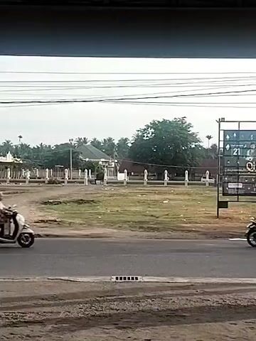 A short video clip shows a scooter with a rider moving from right to left across the frame. The rider is wearing a light-colored hijab. The scooter is white and has a dark seat. The rider is partially obscured by their clothing. In the background, there is a green field with a row of white posts and a building with a green roof. To the right of the field is a large billboard. Power lines are visible across the top of the frame. The road in the foreground is asphalt.