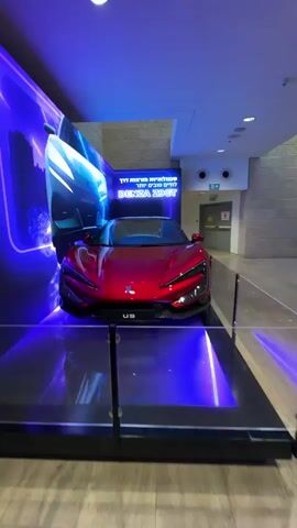 The video shows a red sports car with the badge U9 on the front, on display. The car is positioned on a platform with blue lighting. Behind the car is a large screen displaying images of other vehicles, including a light blue sports car from the rear and a white hatchback from the side, against a backdrop of a cityscape. The display area appears to be indoors, possibly in a showroom or at an event, with transparent barriers around the car and a glimpse of an exit sign in the background. The camera pans slightly, giving a rotating view of the red car.