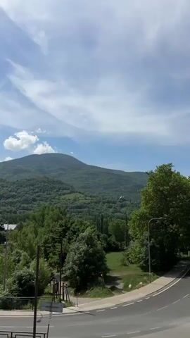 The video shows a helicopter flying over a mountainous, forested landscape. The helicopter, identified as "Guardia Civil," flies from the right side of the frame towards the camera. The background includes a large, forested mountain range with a partly cloudy sky above. The foreground has trees and a portion of a road with street lights. The camera follows the helicopter's movement.