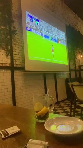 A video shows a sports game being displayed on a projector screen in what appears to be a restaurant or bar setting. The game is soccer, with players visible in their uniforms on the green field displayed on the screen. The screen also shows score information and advertisements. In the foreground, on a wooden table, there is a clear glass mug containing liquid and slices of fruit, possibly lemon. There is also a small white saucer and packets of sugar or sweetener on the table. The background includes a wall with white tiles and faux vines or greenery. The floor has a black and white checkered pattern. There is a chair visible to the right of the table.