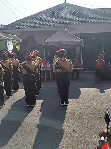 A group of young people in matching tan and brown uniforms, some wearing red hats, are lined up in formation on a paved street. They are performing a synchronized dance or drill, with their arms extended. Some are looking forward, others are looking to the side. A person on a scooter is in the foreground, facing away from the camera, obscuring the view of some of the people. In the background, there is a building with a tiled roof and a white wall. There is also a canopy with a table and chairs set up. Sunlight is casting long shadows on the street.