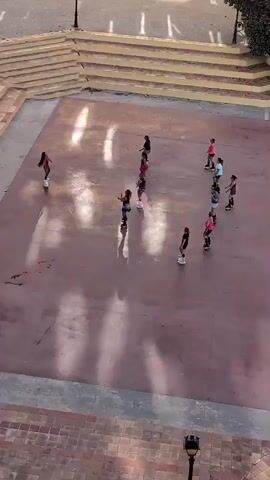 A group of young girls on roller skates are arranged in a circular pattern on a reddish-brown outdoor paved area. The area appears to be a courtyard or plaza with tiered seating in the background. The girls are mostly facing inward towards the center of the circle. The lighting suggests it is daytime, with shadows cast on the ground. The video cuts to a marina scene with docked boats and colorful buildings in the background.