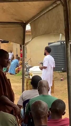 The video shows an outdoor gathering under a tent, with a man in a white shirt and pants standing at a makeshift podium. He is holding a small object in his left hand and gesturing with his right. In front of him, a group of people, mostly older adults, are seated, facing him. Behind the speaker, a white coffin or casket is visible on the podium, along with some other items. To the right of the podium, a large black water tank is situated near a building. The environment appears to be a rural or semi-rural setting, with dirt ground and simple structures in the background. Several other individuals are visible in the foreground, their backs to the camera, also seated and facing the speaker. A few people are standing in the background on the left side of the frame, observing the proceedings. The overall scene suggests a formal gathering, possibly a funeral or religious service.