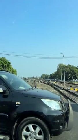 The video shows a view from inside a vehicle traveling on a road that intersects with railroad tracks. The scene is illuminated by bright daylight under a clear blue sky.

At the beginning of the video, the side of a black SUV is visible, with tinted windows and a silver roof rack.

As the vehicle moves, it approaches a railway crossing. A man wearing a blue shirt and a hat is seen pushing a bicycle with a passenger carriage attached to it across the tracks.

Further along, a black flatbed truck is parked or moving slowly near the tracks.

Next, a white van is positioned at the railway crossing.

Finally, an elderly man wearing a brown vest over a greenish shirt and grey pants is walking away from the camera, carrying a white plastic bag in his left hand. He appears to be walking alongside the white van, which is partially obscuring him from view. The background consists of trees and railway infrastructure, including multiple sets of tracks and overhead wires.