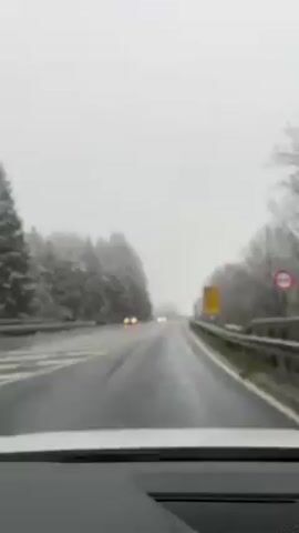 The view from inside a car shows a gray day with snow or sleet on the ground, trees and roadside objects. Rain is falling, obscuring visibility. The wipers of the car are active. Two cars are driving in the distance. The road surface is wet. The guardrail on the right side of the road is metal.  A yellow sign is visible on the right.