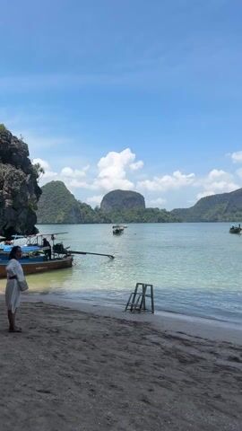 The video shows a beach scene in Thailand with a variety of boats in the water. The first shot depicts a longtail boat near the shore, with several more boats in the distance against a backdrop of islands and a partly cloudy sky. The camera then pans right, showing additional boats including speedboats docked at the beach. People can be seen on some of the boats. The final shot moves under a rock overhang, framing a view of the boats and islands. The color of the water is a light aquamarine.