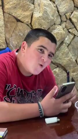 The video shows a close-up of a young person wearing a red t-shirt. The person has dark hair and is looking at a smartphone. The background appears to be a stone wall. The person's facial expressions change throughout the video, showing surprise, mild exasperation, and then a slight smile. The person's left hand is visible, holding the phone. A black bracelet is visible on the person's left wrist.